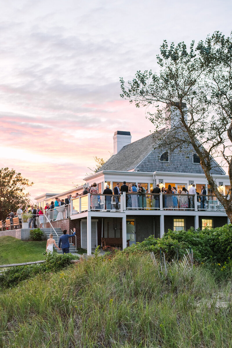 bride and groom waving at guests on the deck at sunset during their chatham beach and tennis club wedding in chatham massachusetts