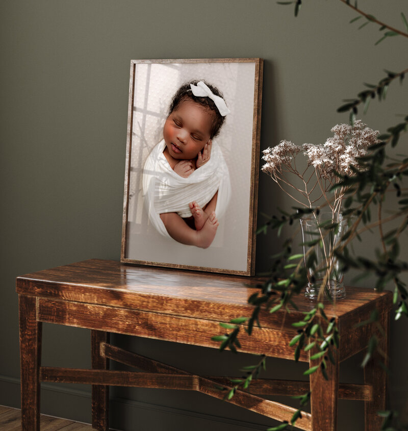 A framed photo of a peacefully sleeping baby, swaddled in white, leans against a dark green wall. This serene moment, captured by a Charlotte newborn photographer, rests on a rustic wooden table with dried flowers in a glass vase nearby. Soft lighting enhances the tranquil atmosphere.