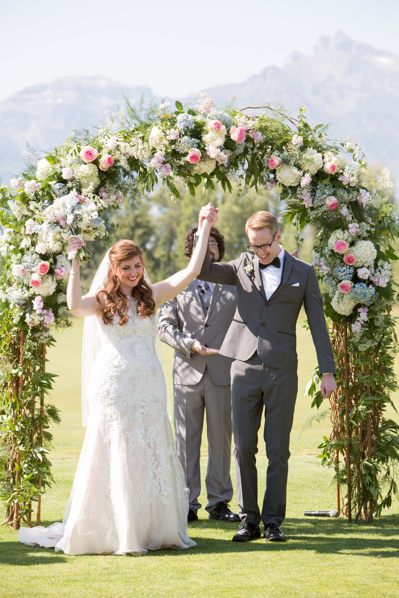A newlywed couple celebrates at a Jackson Hole Golf & Tennis Club wedding, standing under a stunning floral arch with the majestic Tetons in the background. The bride joyfully raises her bouquet, holding hands with her groom in a blissful post-ceremony moment.