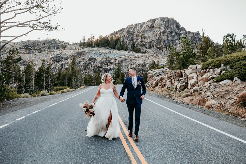 A joyful bride and groom, holding hands and smiling widely, run together down a scenic mountain road in Lake Tahoe, surrounded by rugged granite cliffs and evergreen trees.