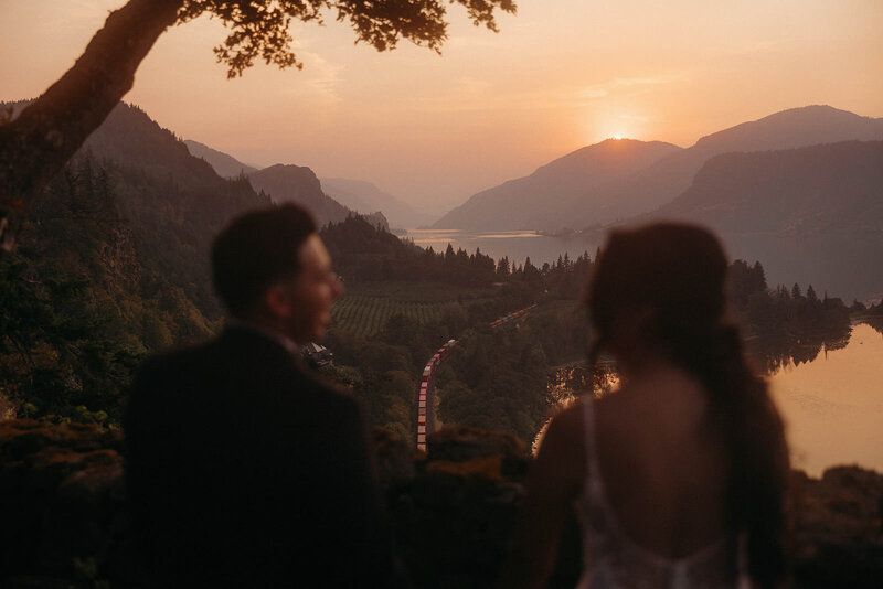Bride and groom watching the sunset over the Columbia River Gorge, with a winding train below, captured by a PNW wedding photographer.