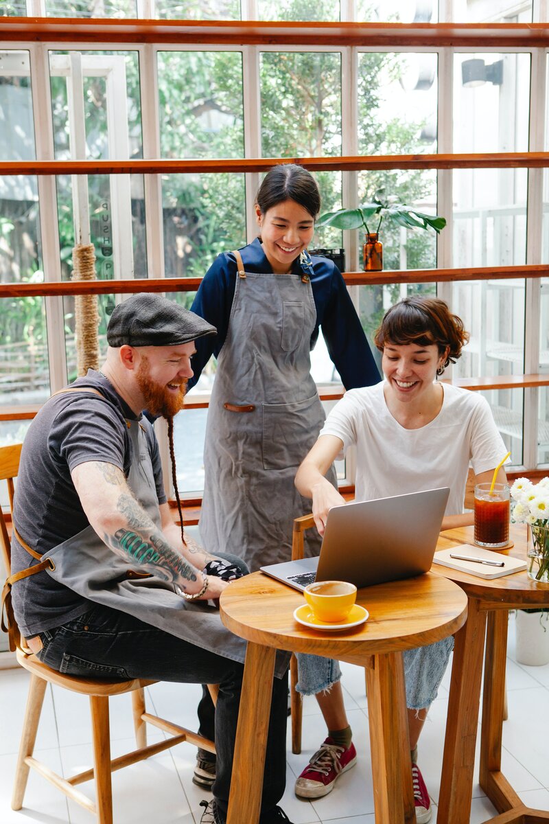 This is an image of three people gathered around a computer and smiling. One is a male presenting person and two are female presenting. They appear to be in a coffee shop.
