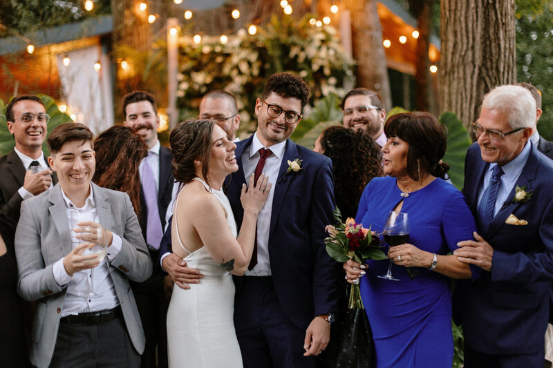 A couple poses closely at a festive outdoor event, surrounded by well-dressed guests, warmly lit with string lights and greenery.