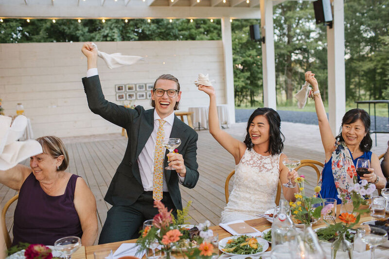 Catskills wedding couple smile big as they sit at floral-covered table and raise their glasses to a toast.