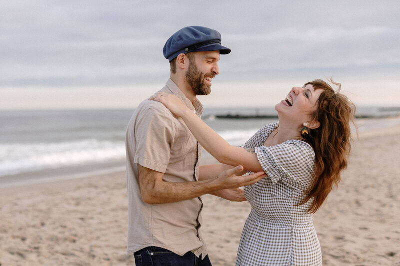 A couple shares a joyful moment on the beach, with soft waves and a cloudy sky in the background, conveying affection and connection.