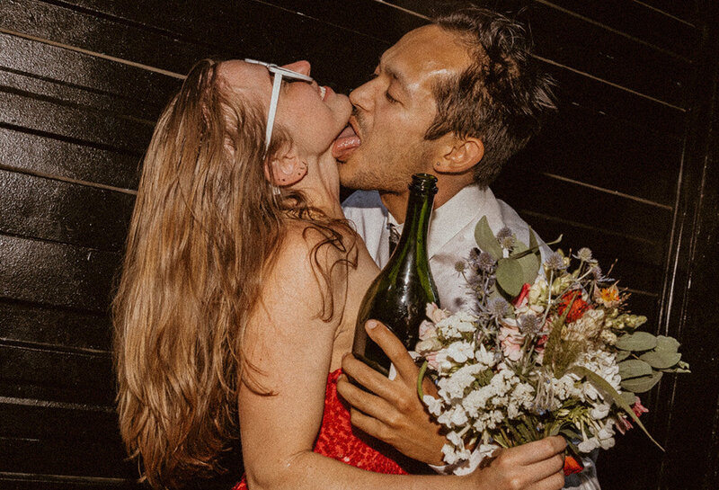 groom licks champagne off bride wearing red dress in a grunge alley 