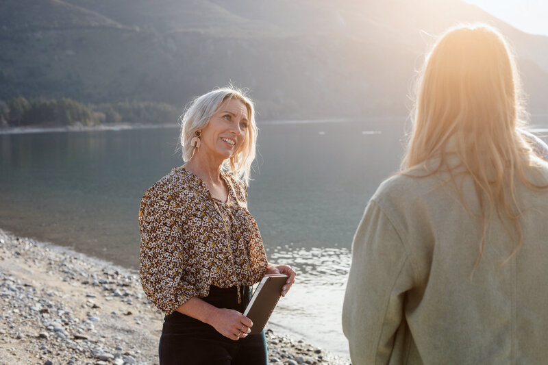 Kirsty and the couple are standing on the shores of a misty and very still lake. Kirsty is facing the couple and has her head tilted to the side like she is engaged in conversation with them. The couple are blurred and not really in the image.