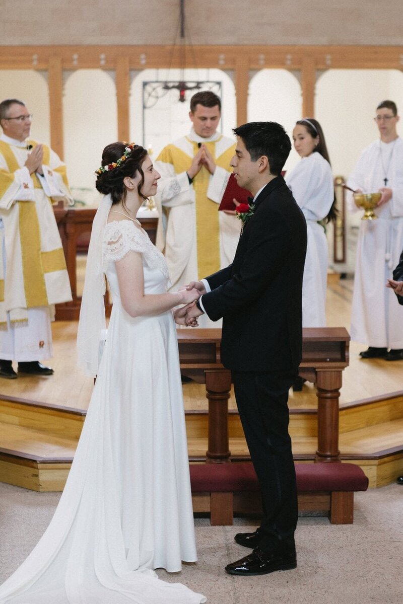 Bride and groom make their vows in front of a priest and altar servers.
