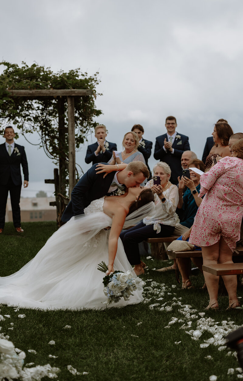 A bride in a white dress and groom in a suit do a dip-kiss in the aisle after their wedding ceremony holding a colorful bouquet on a family-lined path. Sunlight filters through the clouds, casting a warm glow, capturing the essence of fine art weddings and portraits in New England.