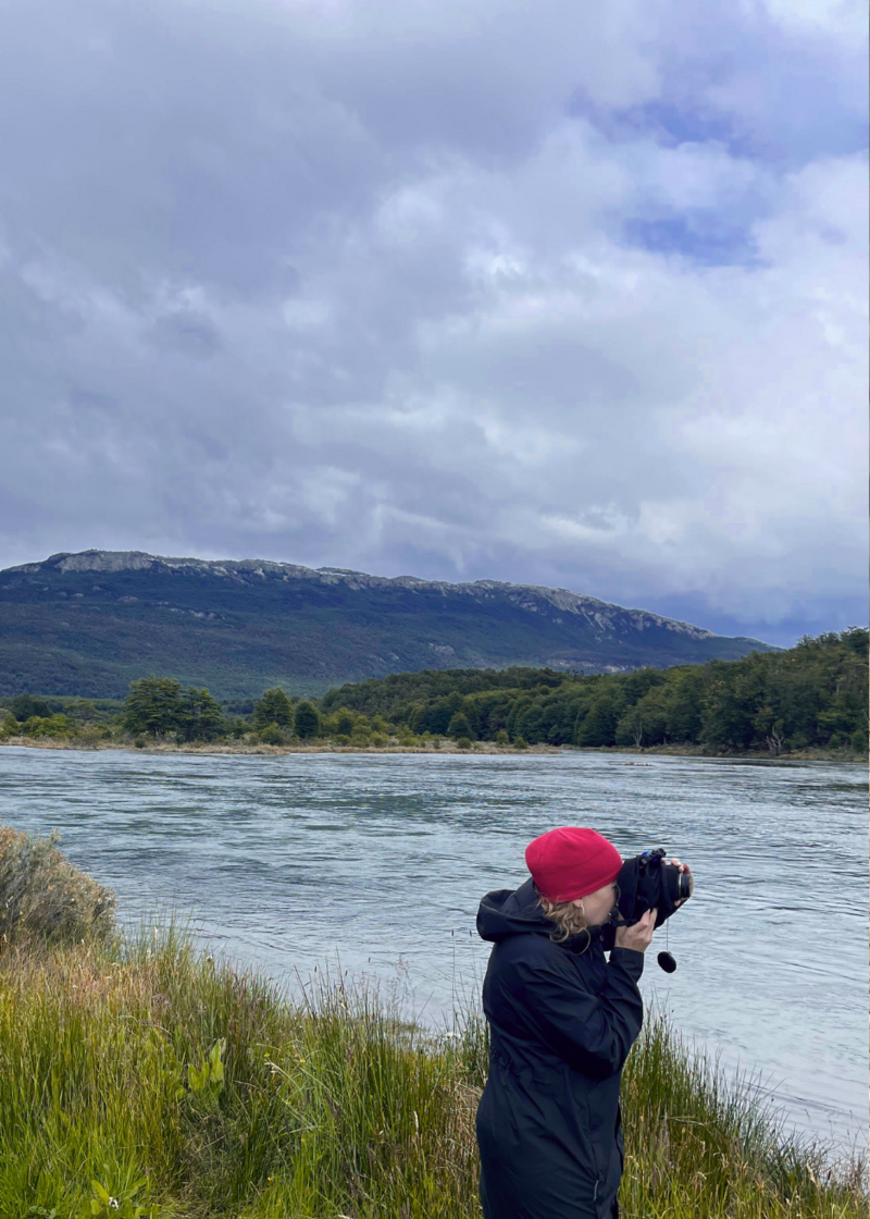 Alicia is standing next to a river in the process of taking a photo, wearing a black rain jacket and red hat.