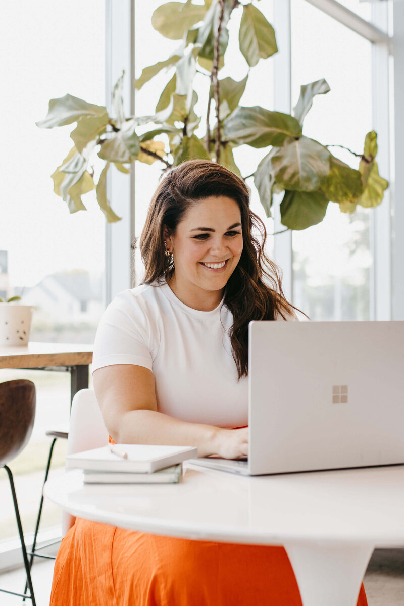 woman with dark hair wearing white shirt and orange skirt on her laptop at a table