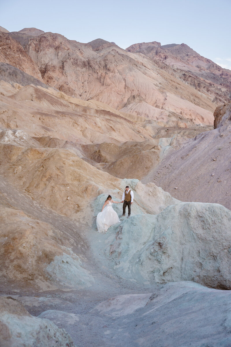 A couple in wedding clothes hikes over colorful hills in the Death Valley desert