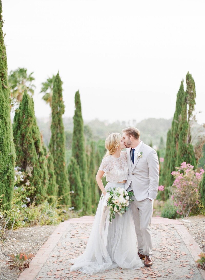 Kohnur couple kissing in a Mediterranean garden with cypress trees. Bride in a two-piece lace/tulle gown and textured low updo. Tuscany inspired Wedding at Southern California