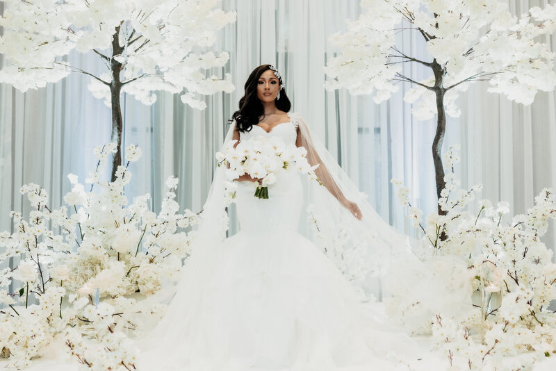 Bride in an elegant mermaid-style gown holding cascading white orchids, standing beneath a floral canopy of white cherry blossom trees and ivory blooms, designed by Wells Design Co.