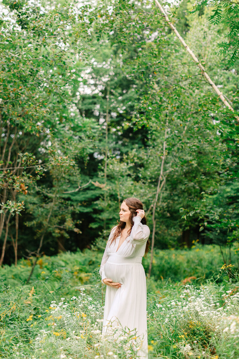 Pregnant woman with brown hair in a white dress cradling her belly with one hand looking to the left in the lush green woods