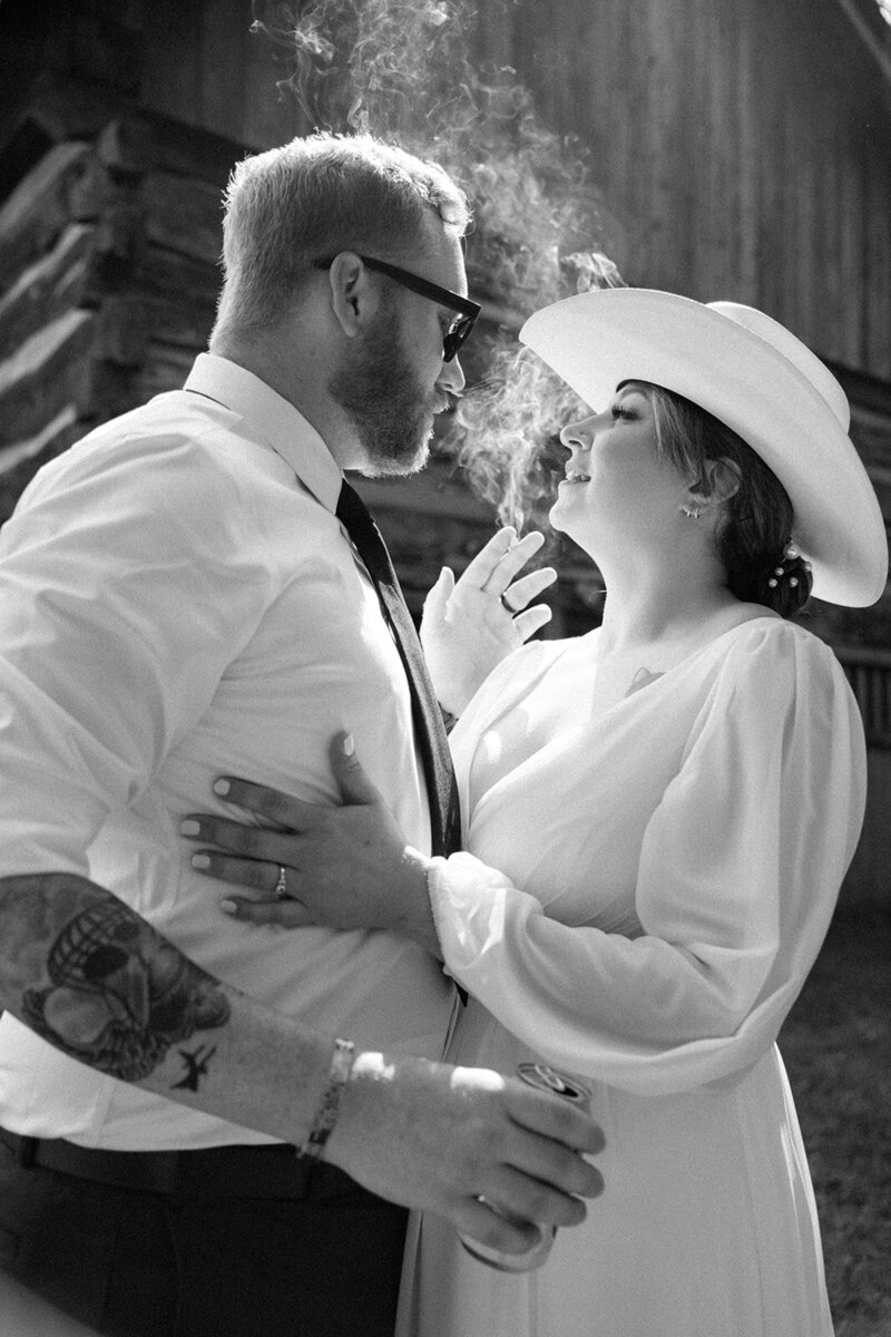 A bride in white sports a white cowboy had and looks into her husbands face while smoke from his cigarette smolders behind them.