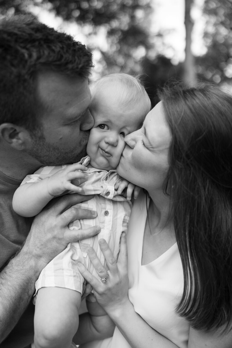 Mom and dad squish baby in the middle and kiss him on the cheek