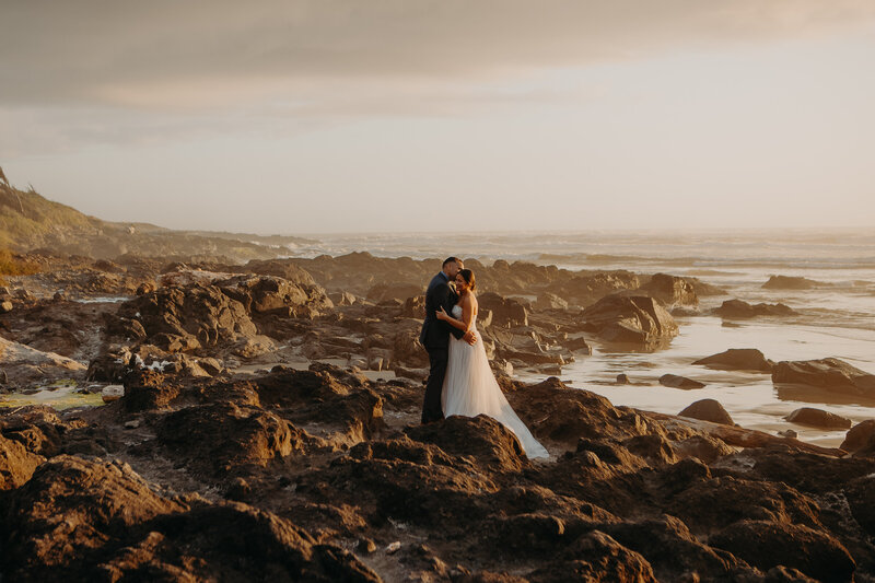a couple at rockaway beach stand together in wedding outfits on a rocky shore with orange sunset lighting