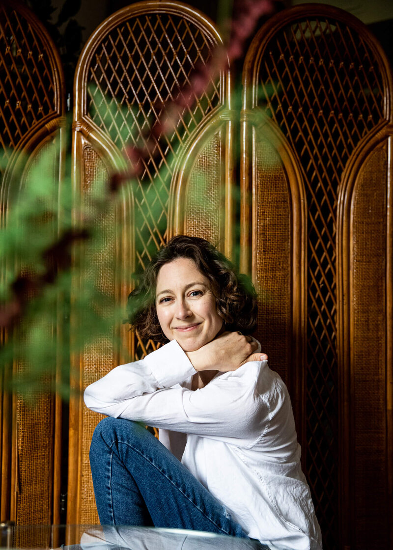 Smiling image of photographer of AM Family Photography shot through  plant leaves, lit by a nearby window