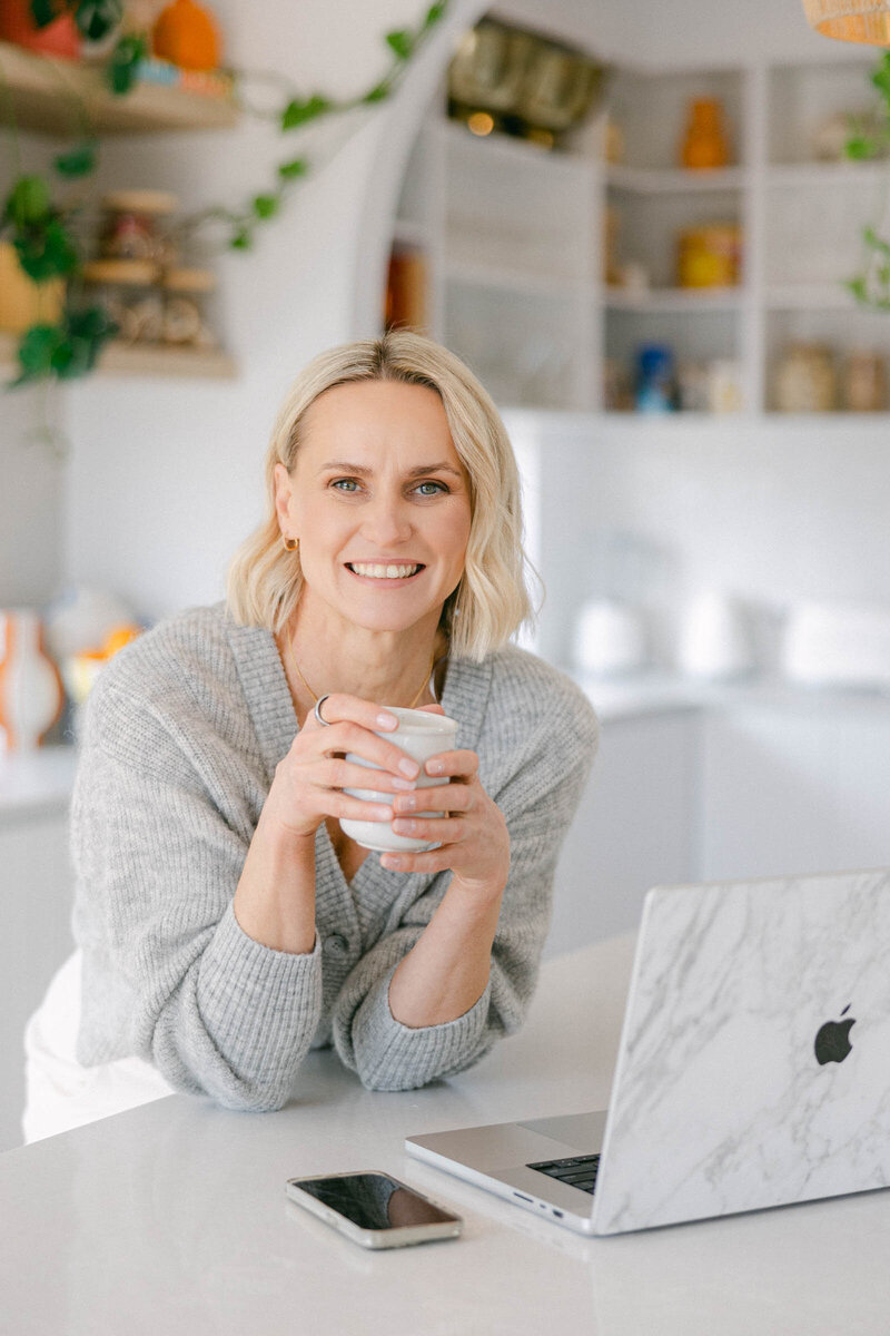 Zelda Green, money and mindset educator and entrepreneur, relaxed and smiling in her kitchen with a cup of tea while working on her profit building business on her laptop