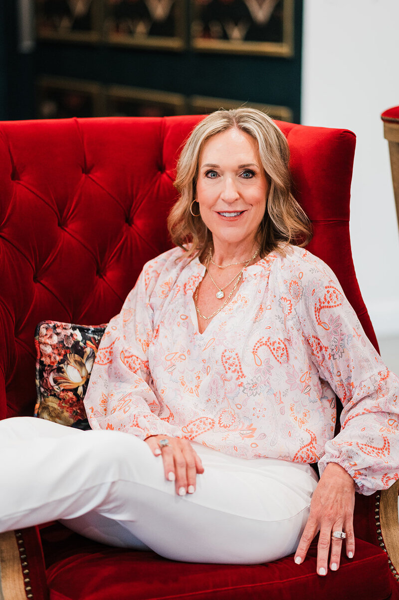 A wedding planner sits on a red velvet chair, smiling warmly, showcasing the welcoming and personable style of Socially Significant Events’ planning team.