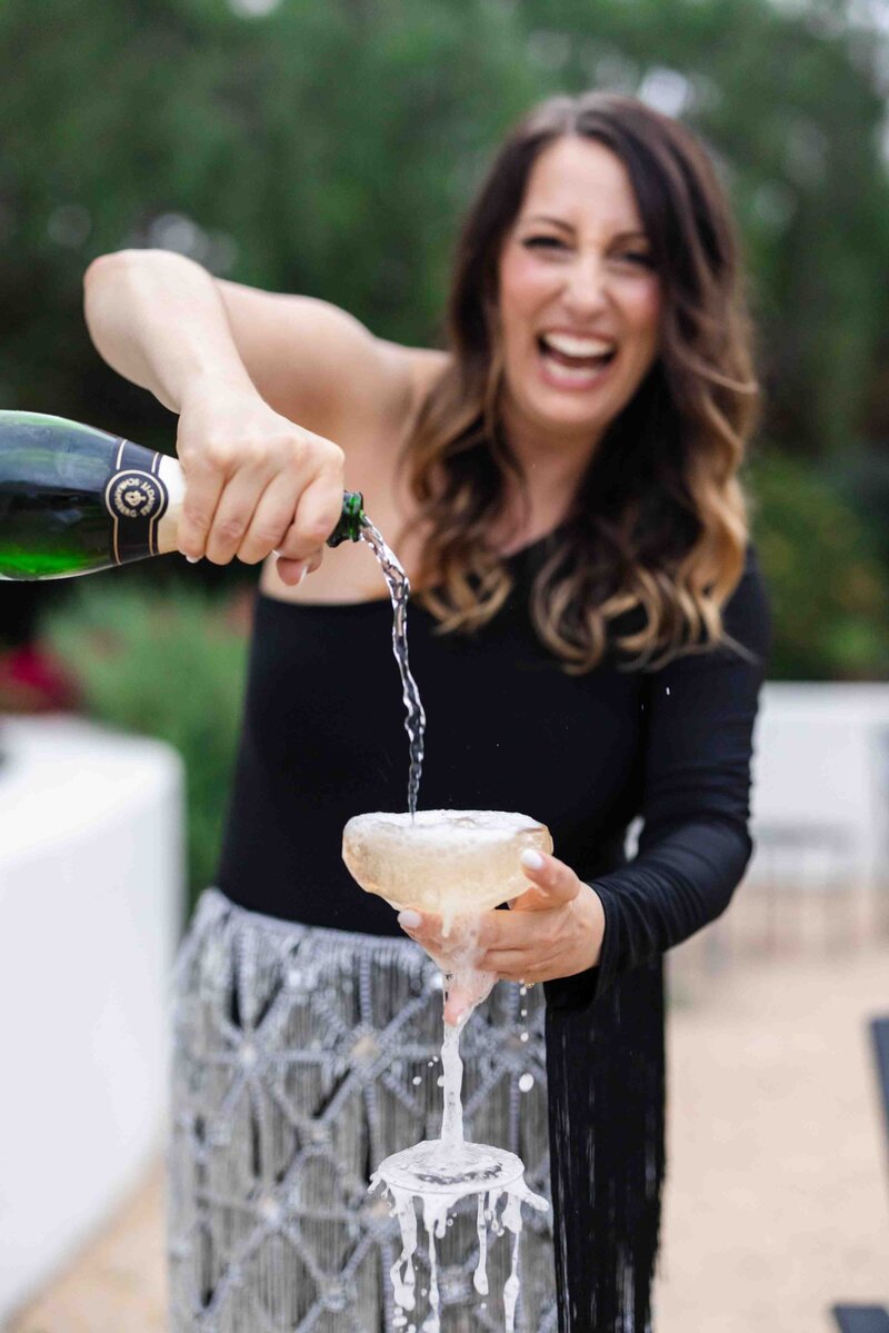 Candid celebration moment capturing joyful laughter while pouring champagne into vintage coupe glass in outdoor garden setting