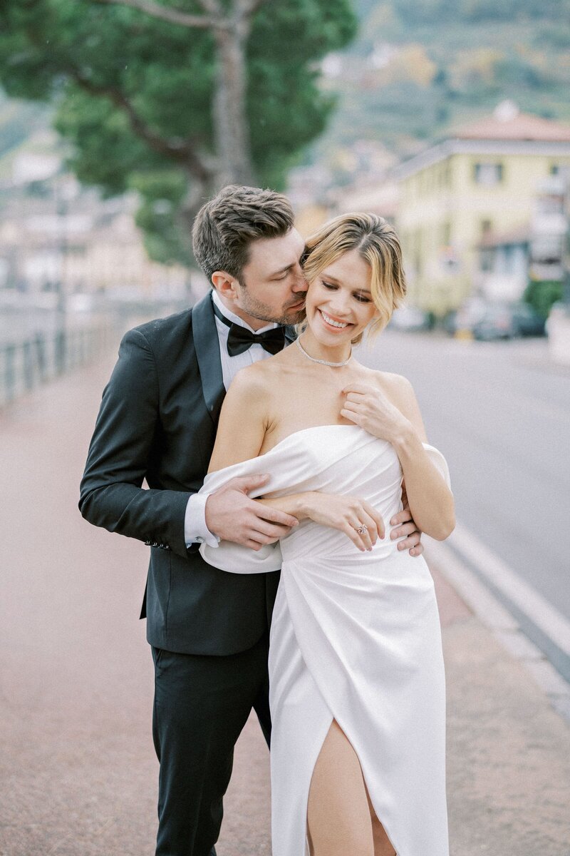 groom-kisses-bride-cheek-while-hugging-her-from-while-standing-shore-lake