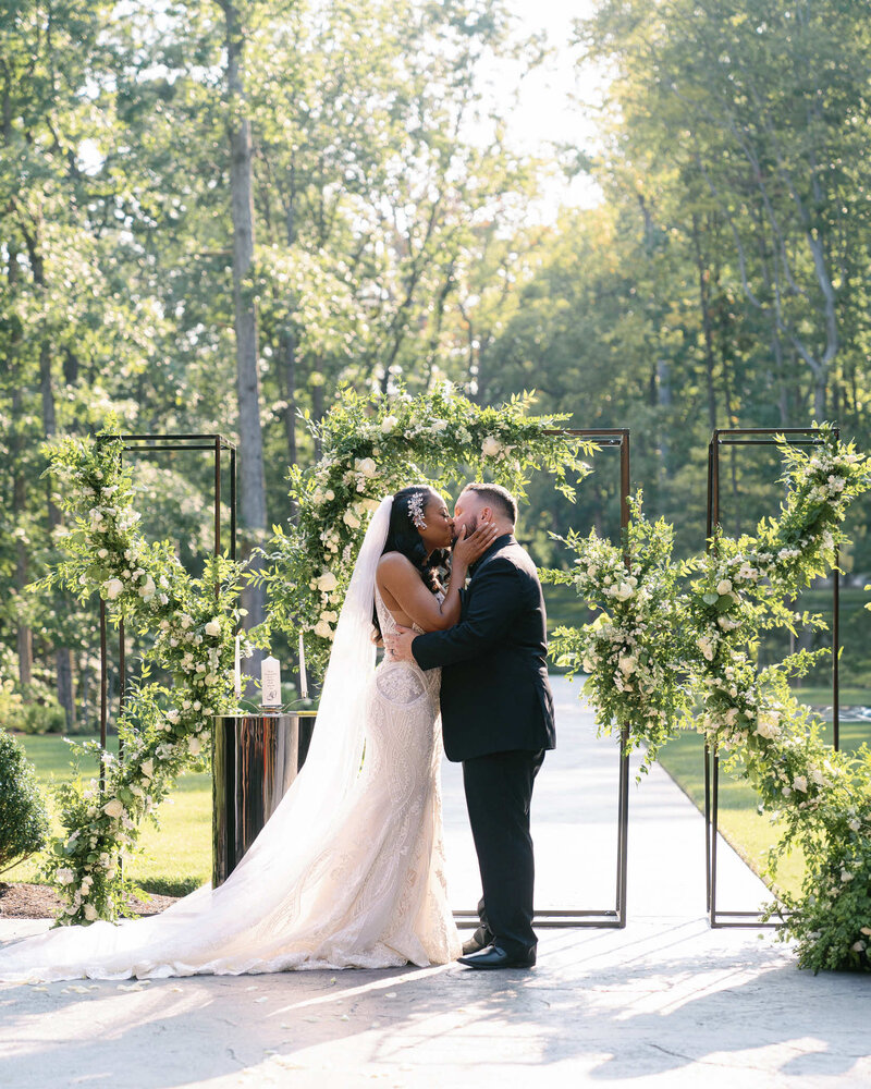 Couple kissing after their wedding ceremony in a lush garden area surrounded by trees and ivy with a iron gate.