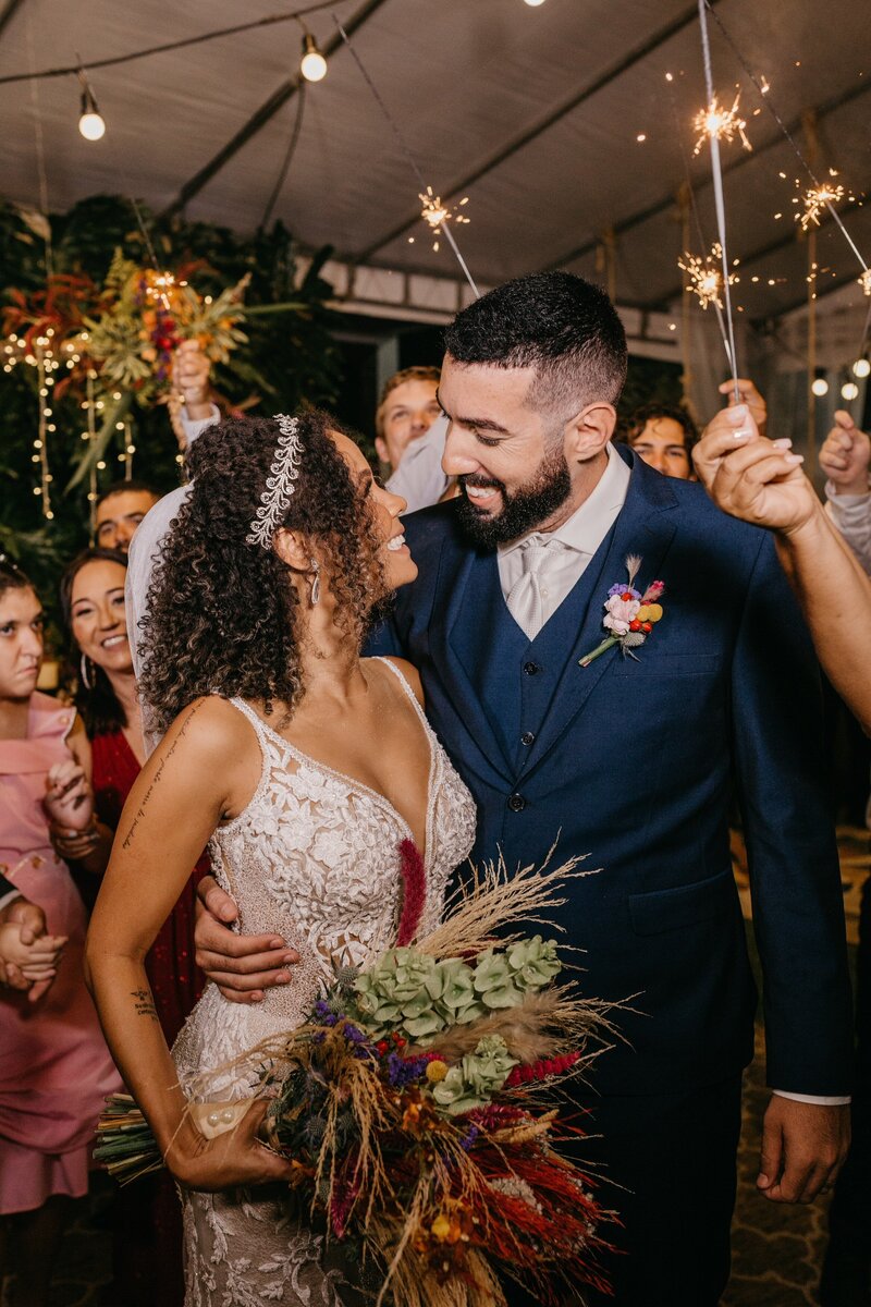 A bride and groom looking at each other smiling while their guests hold sparklers around them.