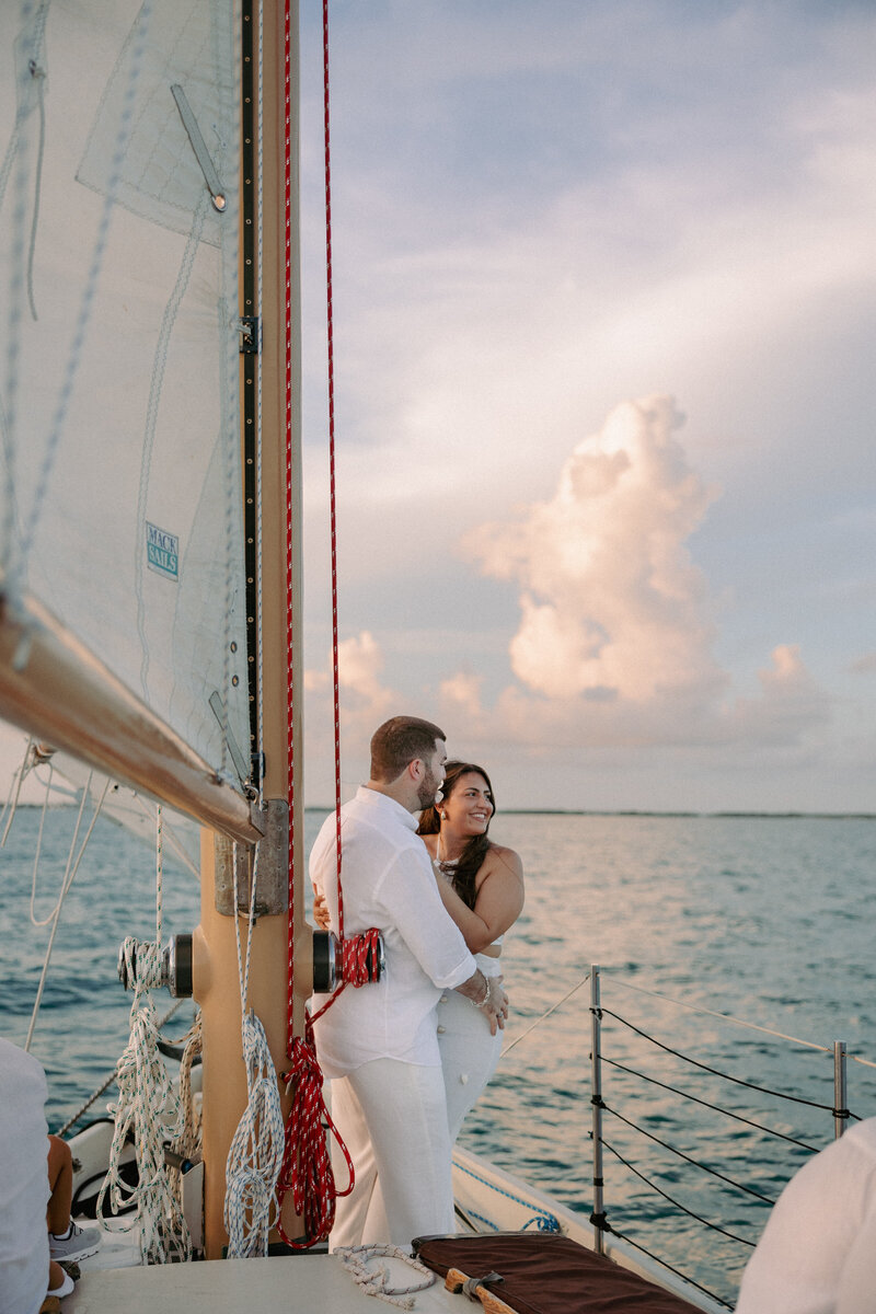 bride and groom embracing on the beach with the ocean and a sailboat in the background