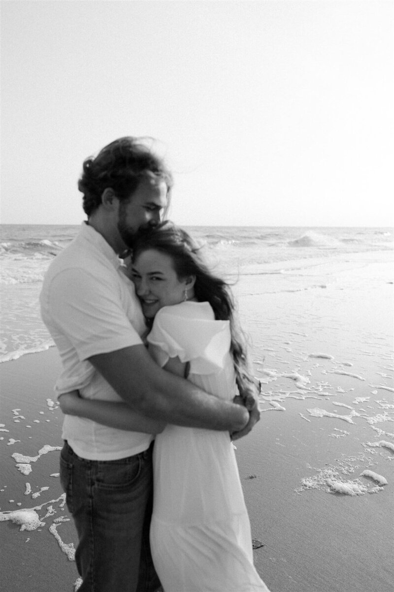 A happy couple embraces on a windy beach at sunset in black and white