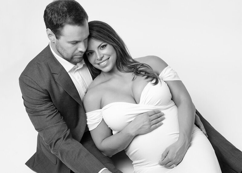 husband with pregnant wife leaning against him wearing white maternity gown