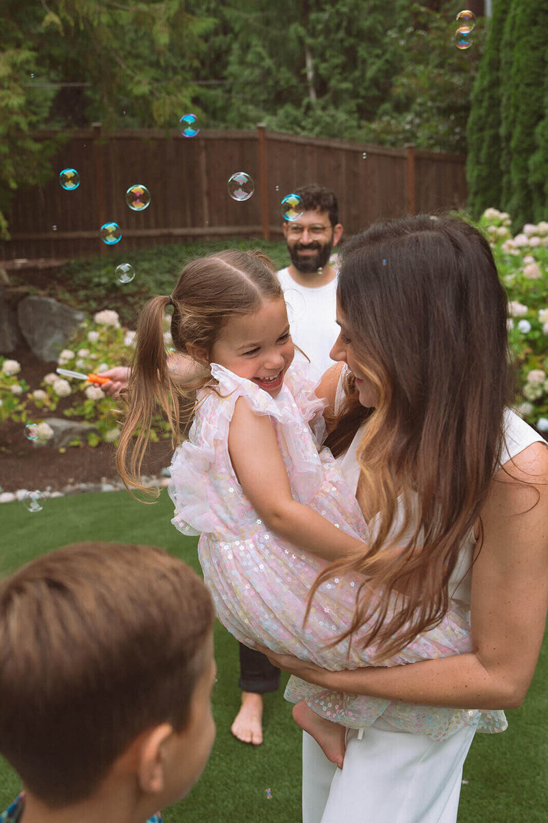female holding little girl in pink dress with family at home in backyard with bubbles