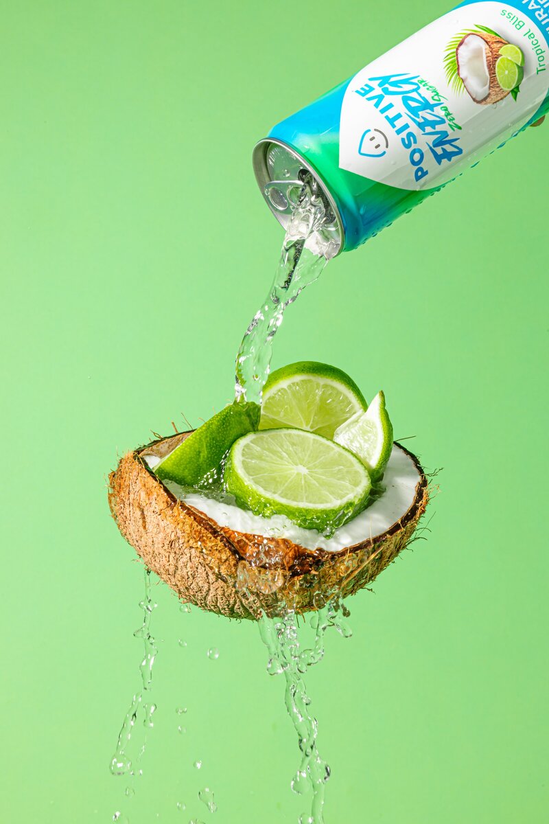 A can of coconut water being poured into a green coconut with lime wedges on a green background. The can has a blue and green label with white text that says "Positive" on top and "Energy" below it.