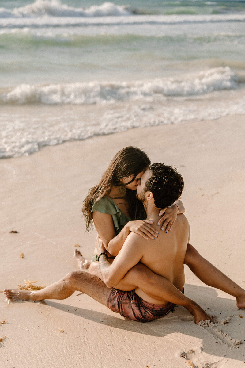 Engaged couple sitting on the sand at a Mexico beach, embracing by the shoreline during golden hour.