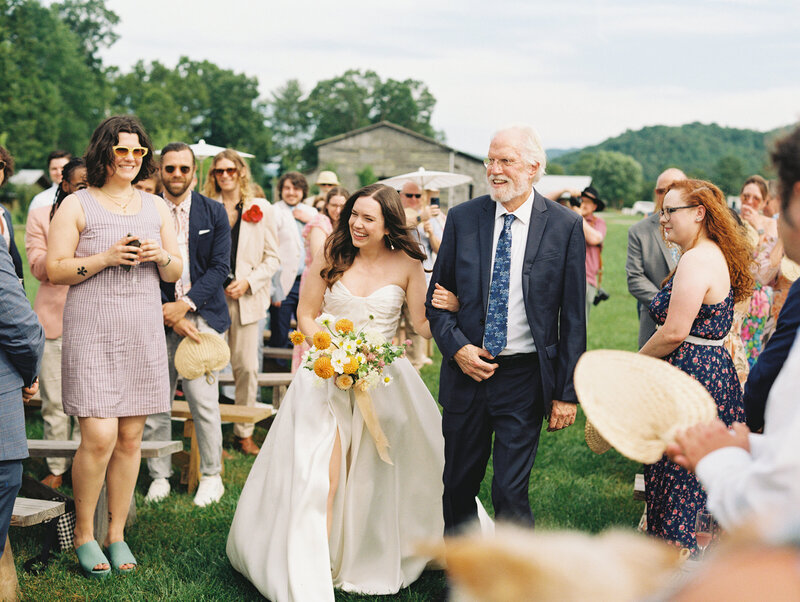 A beautiful and beaming bride is escorted by her father next to the joyful and colorful wedding guests during this summer wedding in North Carolina, by film photographer Megan Lynn of My Sun and Stars Co.