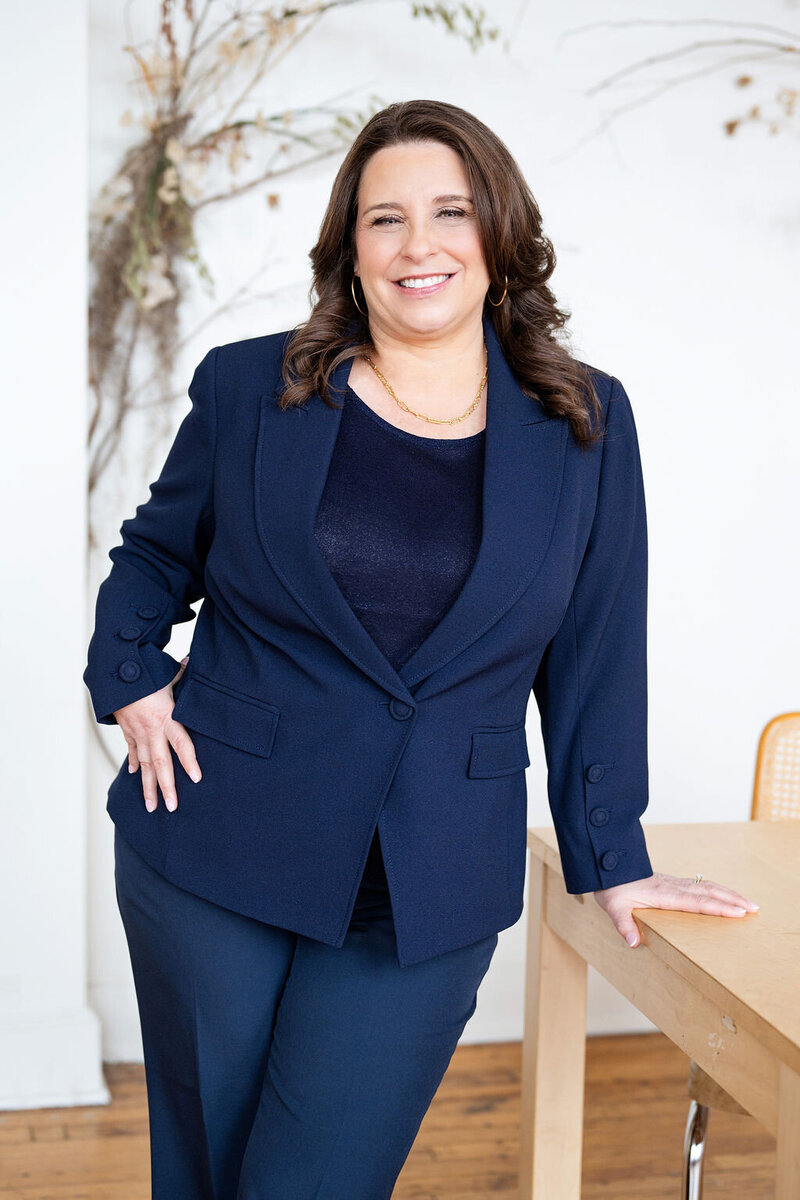 Chris standing confidently in a navy suit beside a desk with flowers and books, smiling at the camera.