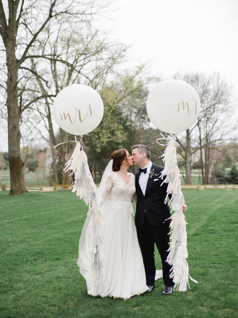 Bride in lace gown and groom in black tuxedo kissing holding mr. & mrs. balloons with tasseled fringe on the grass at Cornman Farms