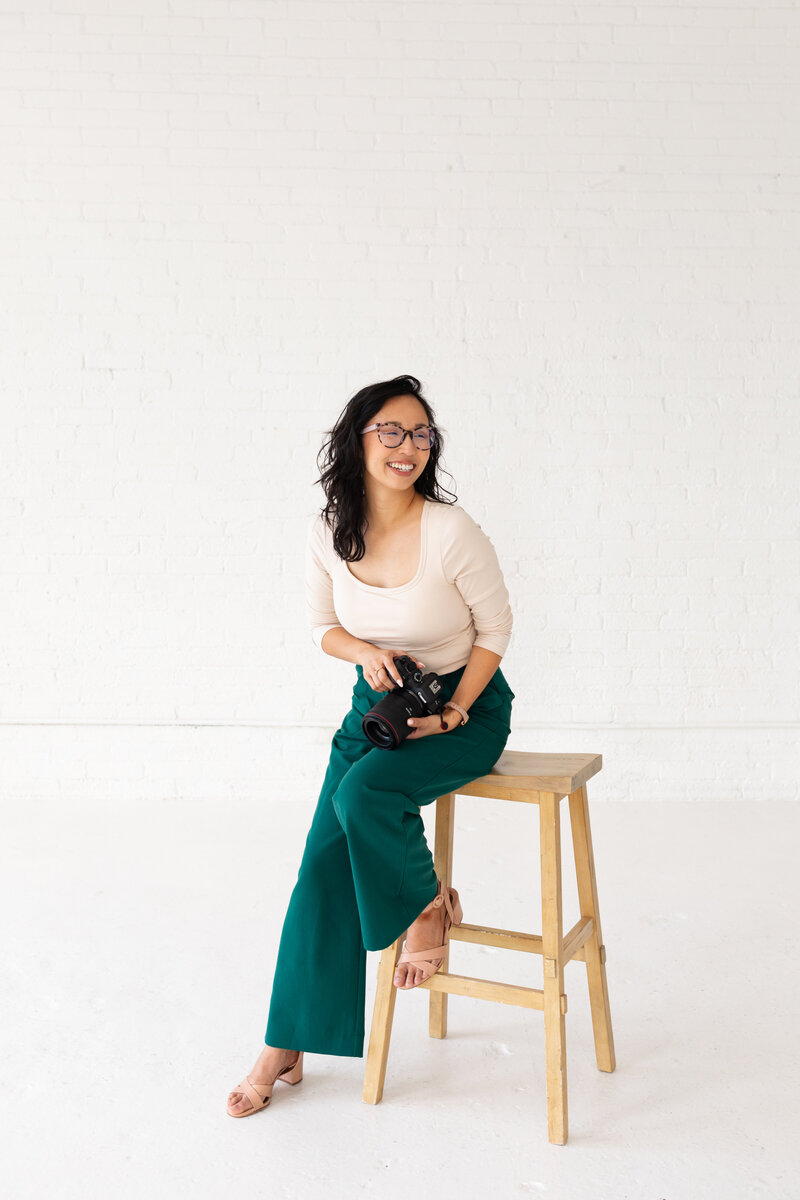 Anh Nguyen, portrait photographer sitting on a stool with a camera.