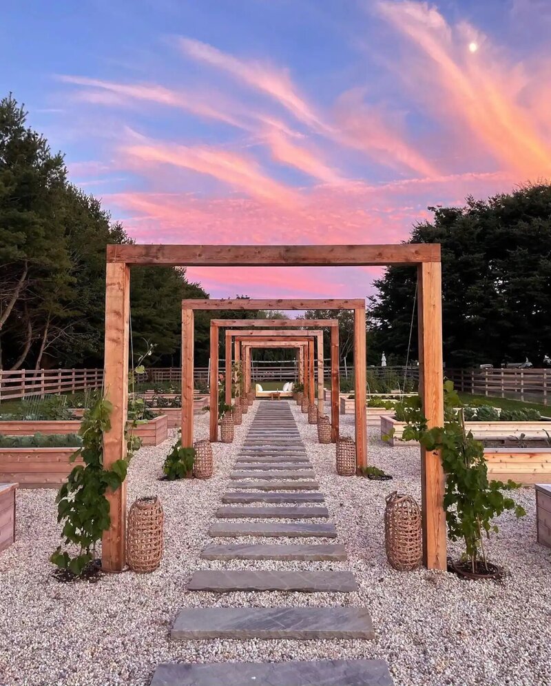 Sunset photo of a row of rectangular wooden trellises over a stone walkway in the garden of this venue