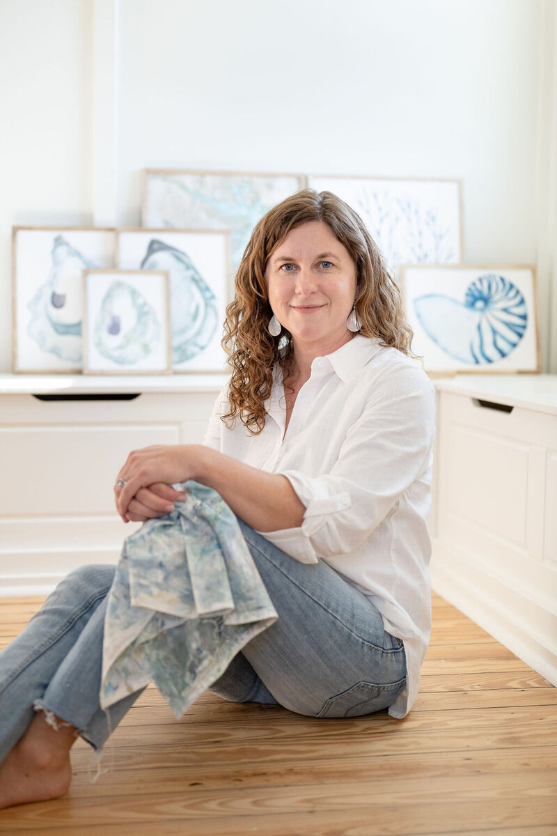 watercolor artist wearing a white button up and light jeans sitting on a wooden floor holding a tea towel with her coastal artwork displayed in the background in wilmington, north carolina