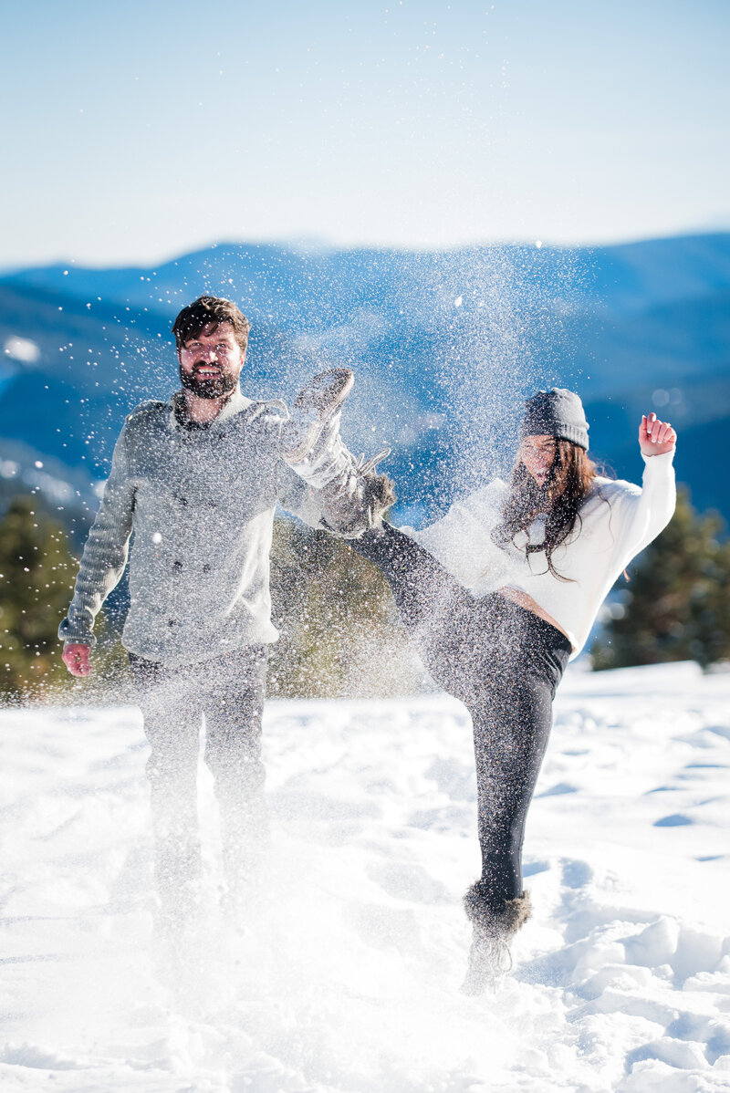 A couple stands on a mountainside and kicks snow toward the camera during their engagement session.