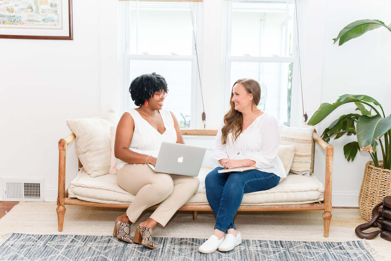 two women sitting on a couch in front of a windown smiling at each other while doing workflow design.