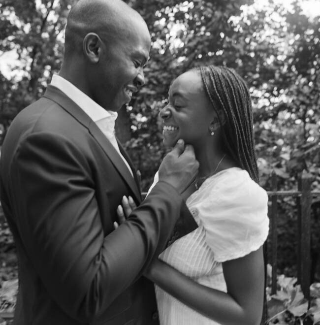 A couple shares a joyful moment, smiling at each other in a leafy outdoor setting.