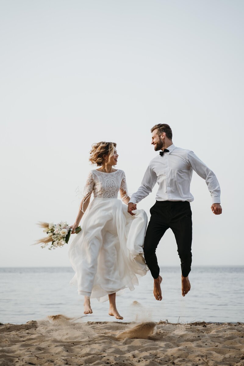 Mariés se tenant la main et se regardant dans les yeux faisant un saut ensemble sur la plage. Organisation de mariage proposée par Nos moments Ensemble, agence évènementielle Nantes Loire Atlantique Vendée