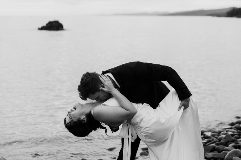 Married couple taking portraits in Grand Marais, MN, captured in black and white. The groom dips the bride as she laughs, holding his neck, with her knee propped up near his hip, during a wedding photography session in MN.