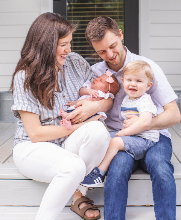 a family with mother, father, toddler, and baby sitting and smiling on a porch