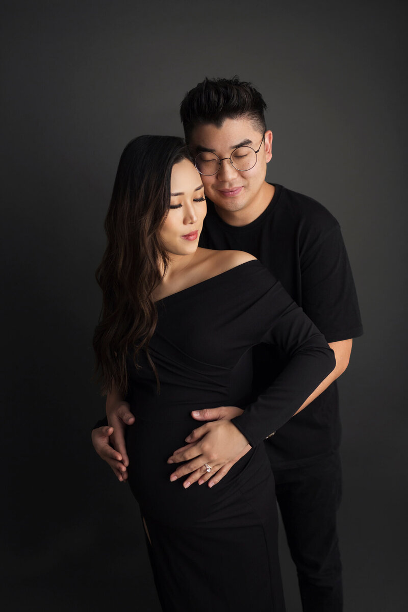 A pregnant woman stands with her partner against a dark background, both wearing black outfits. She is dressed in an off-the-shoulder black dress, and he is wearing a black T-shirt. The partner stands behind her, gently embracing her and cradling her baby bump with both hands. They both have serene expressions, with their eyes closed, conveying a sense of peace and intimacy.