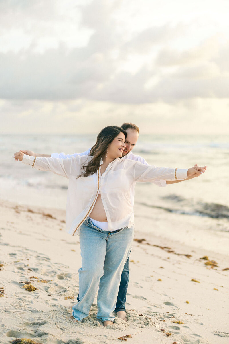 Expecting couple laughing and embracing on the beach during a soft, golden-hour maternity session.