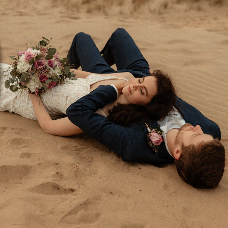 bride and groom laying in the sand snuggling together
